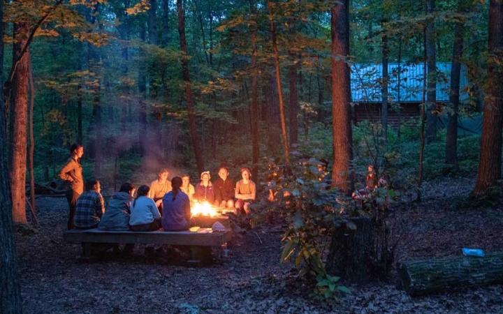 a group of teens sit around a campfire in a wooded area